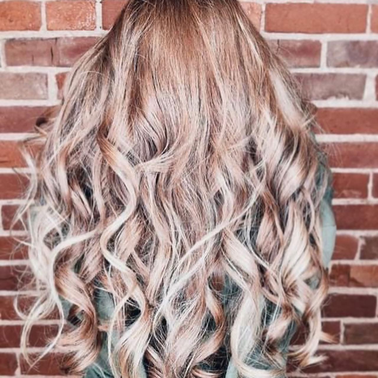 Long wavy blonde balayage with soft caramel ribbons against a warm brick wall